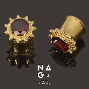 以下が含まれることがあります： 赤い宝石が装飾的な金の枠にセットされた、金製の耳のゲージのペア。ゲージは黒い表面にあります。