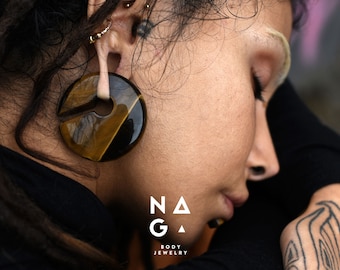 Puede incluir: Primer plano de una persona con un pendiente grande y circular de ojo de tigre. El pendiente es de color marrón oscuro y amarillo. También se muestra el texto "NAGA BODY JEWELRY".