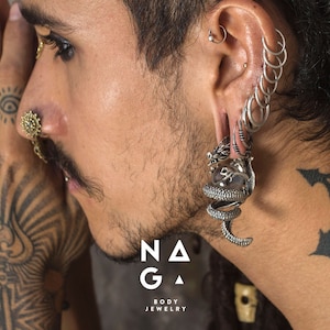 Può includere: Una persona con più piercing alle orecchie indossa un orecchino a forma di drago e serpente in argento. L'orecchino è un gioiello grande e dettagliato. La persona ha anche un anello al naso e tatuaggi sulle mani e sul collo. L'immagine include il testo "NAGA BODY JEWELRY".