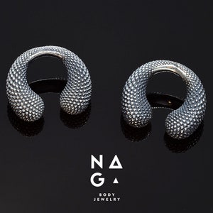 以下が含まれることがあります： 銀製のテクスチャ加工が施された、湾曲したオープンフープイヤリング2つ。イヤリングは耳たぶに着用するように設計されています。