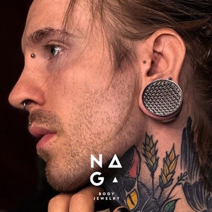 Può includere: Una persona con più piercing, tra cui un piercing al lobo dell'orecchio di grosso calibro con un tappo argentato testurizzato. La persona ha un tatuaggio sul collo di un gatto nero con occhi gialli e spighe di grano.