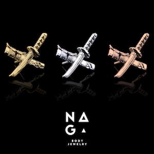 May include: Three miniature crossed katana swords in gold, silver, and rose gold tones are displayed against a black background. The swords are detailed with visible handles and blades. The logo "NAGA BODY JEWELRY" is at the bottom.