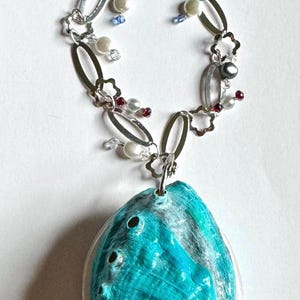 May include: A necklace featuring a large, iridescent turquoise abalone shell pendant suspended from a silver-toned chain. The chain is composed of oval and flower-shaped links, accented with small white pearls, dark grey beads, and tiny red crystals.