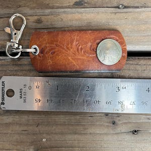 Könnte beinhalten: Ein brauner Leder-Schlüsselanhänger mit einem silbernen Verschluss und einer Nickelmünze. Das Leder hat ein geprägtes Blumenmuster. Ein Metalllineal liegt neben dem Schlüsselanhänger.
