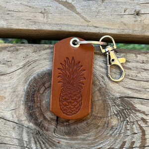 May include: A brown leather keychain with a pineapple design. The keychain has a silver metal ring and a silver metal clasp.