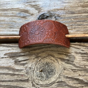 May include: A brown leather hair stick with a floral design. The stick is curved and has a smooth finish.