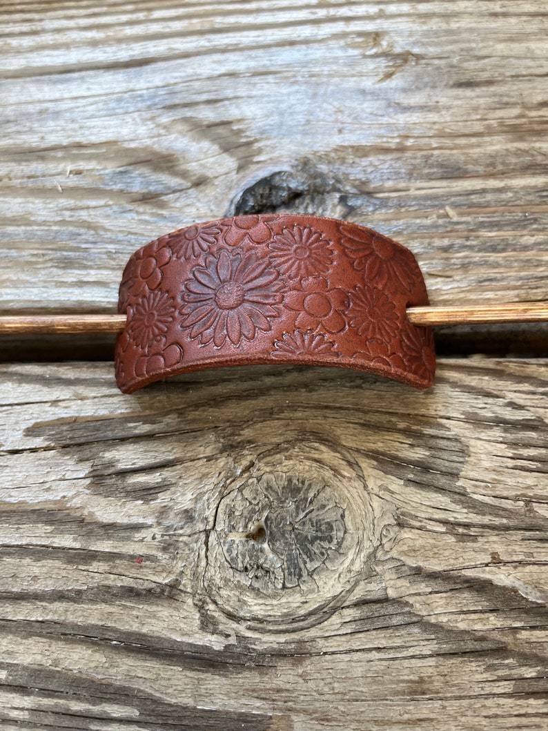 May include: A brown leather hair stick with a floral design. The stick is curved and has a smooth finish.
