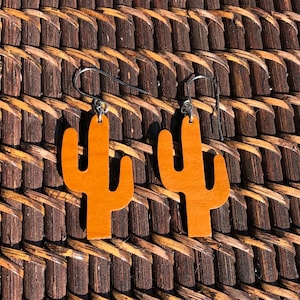 Puede incluir: Un par de pendientes de cactus de cuero marrón con ganchos plateados. Los pendientes tienen forma de cactus simple con un solo tallo y dos brazos.