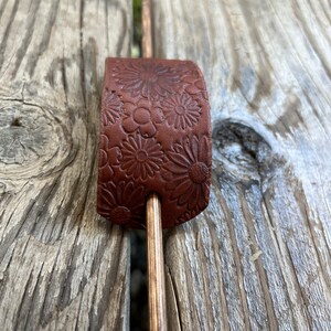May include: A brown leather hair stick with a floral pattern. The stick is made of wood and has a natural finish.