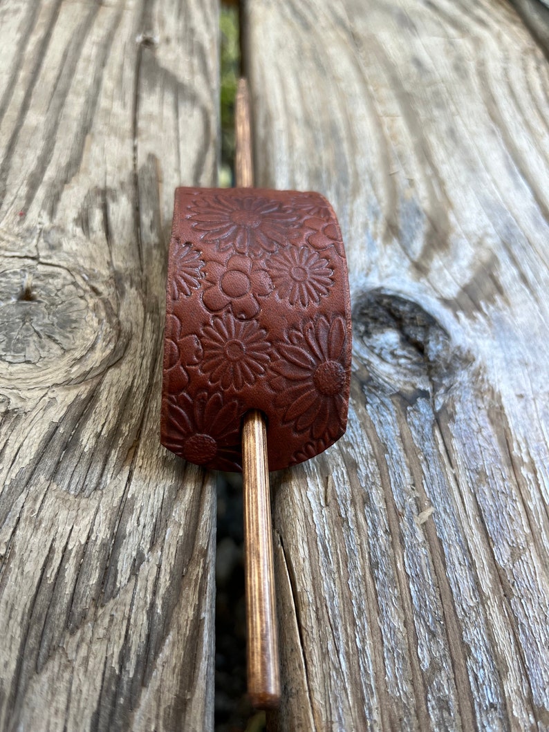 May include: A brown leather hair stick with a floral pattern. The stick is made of wood and has a natural finish.