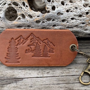 May include: A brown leather keychain with a mountain and trees design. The keychain has a brass key ring.