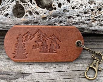 May include: A brown leather keychain with a mountain and trees design. The keychain has a brass key ring.