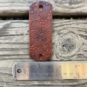 May include: A brown leather strap with a floral design. The strap is approximately 1 inch wide and 2 inches long. The strap has two holes for attaching to a belt or other item.