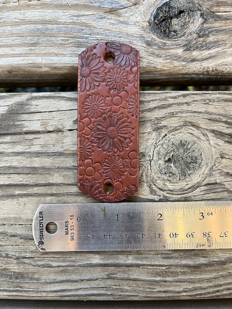 May include: A brown leather strap with a floral design. The strap is approximately 1 inch wide and 2 inches long. The strap has two holes for attaching to a belt or other item.