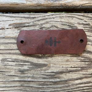 May include: A brown leather strap with two holes and a black logo featuring a tree and a cross.