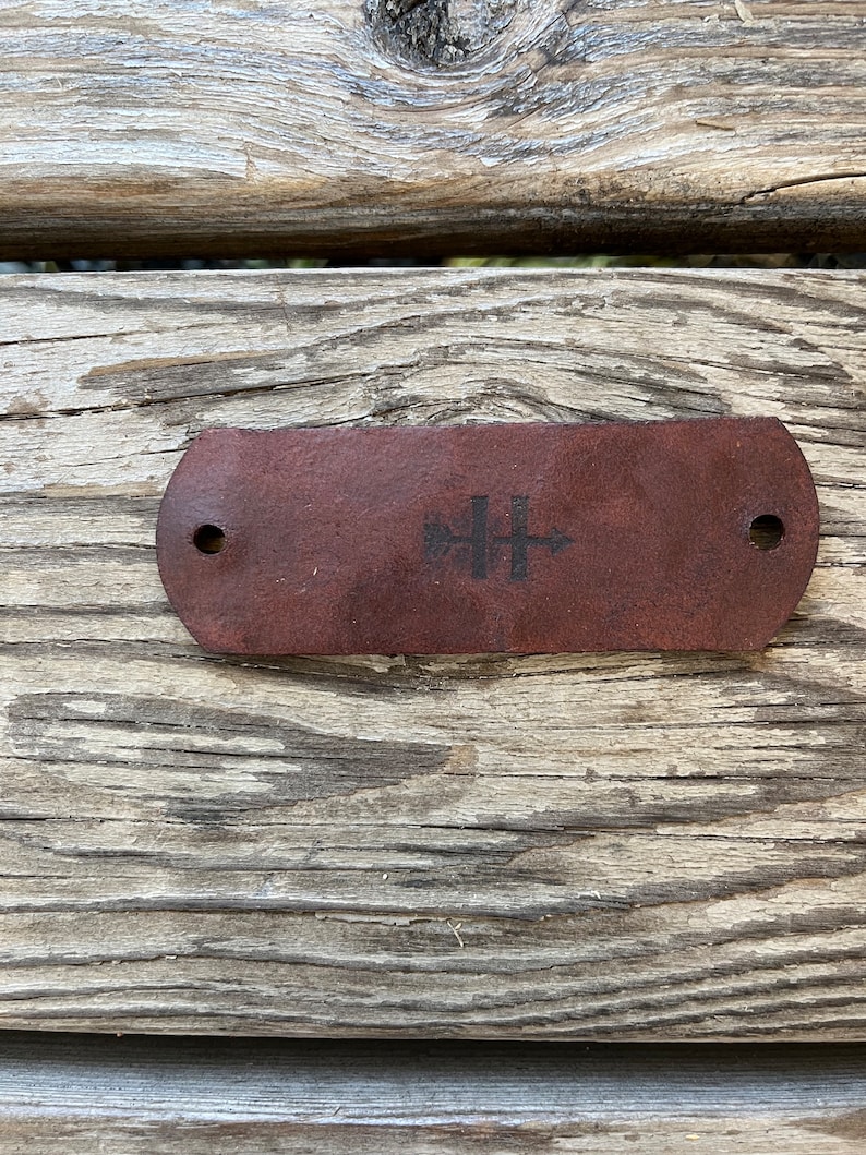 May include: A brown leather strap with two holes and a black logo featuring a tree and a cross.