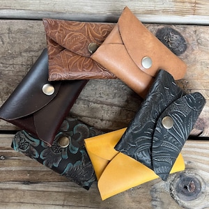 May include: A selection of handmade leather wallets in various colours, including brown, black, and yellow. Some wallets feature embossed floral patterns and secure with metal snap closures. The wallets are arranged on a rustic wooden surface.