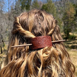 May include: A brown leather hair stick with a floral design. The stick is being used to hold a woman's hair in a bun.