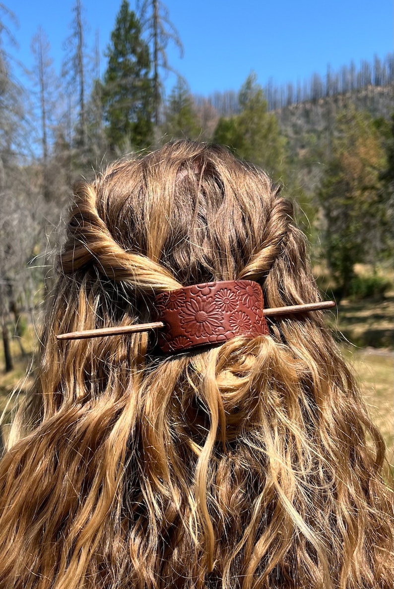 May include: A brown leather hair stick with a floral design. The stick is being used to hold a woman's hair in a bun.