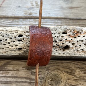 May include: A brown leather hair stick with a floral design. The stick is made of wood and has a simple, elegant design.