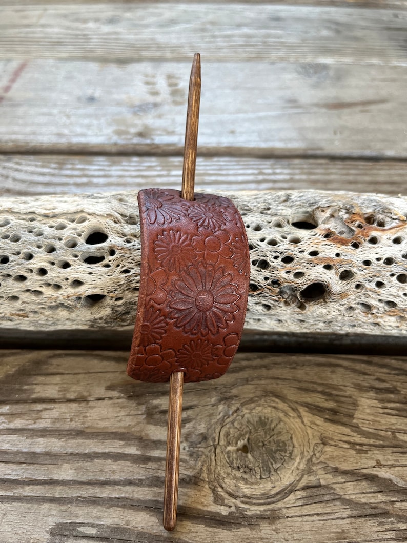 May include: A brown leather hair stick with a floral design. The stick is made of wood and has a simple, elegant design.