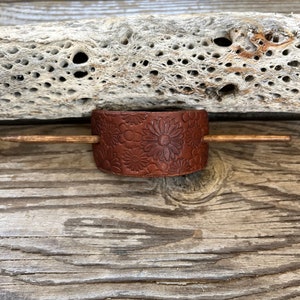 May include: A brown leather hair stick with a floral design. The stick is on a wooden surface.