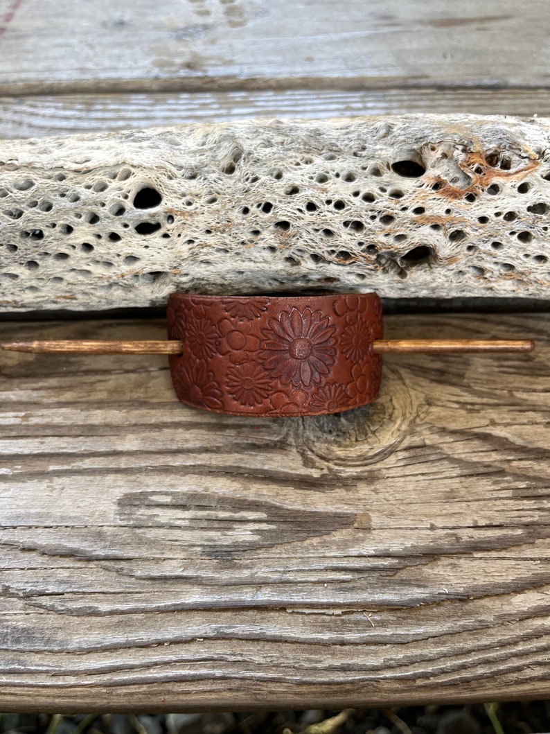 May include: A brown leather hair stick with a floral design. The stick is on a wooden surface.