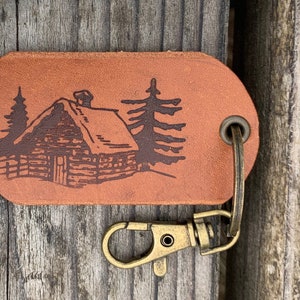 May include: A brown leather keychain with a cabin and trees design. The keychain has a brass key ring and a lobster clasp.
