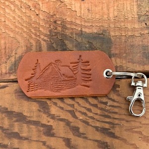 May include: A brown leather keychain with a cabin and trees design. The keychain has a silver metal ring and clasp.
