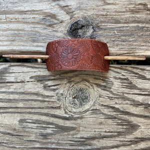 May include: A brown leather hair stick with a floral design. The stick is made of wood and has a brown finish.