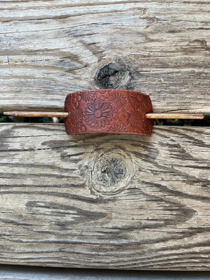 May include: A brown leather hair stick with a floral design. The stick is made of wood and has a brown finish.