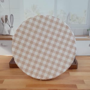 Może przedstawiać: Okrągła osłona na żywność w beżowo-białą kratę. Pokrywa znajduje się na drewnianej tacy, ustawionej na drewnianym blacie. Tło kuchni obejmuje okno, zlew i przybory kuchenne.