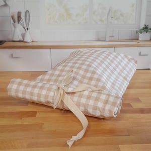 May include: Rolled-up beige and white gingham patterned kitchen mat tied with a matching ribbon. The mat is on a wooden surface, with a kitchen background featuring a window, sink, and cabinets.