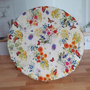 May include: A round, floral-patterned tablecloth featuring a cream background with colorful flowers and butterflies. The flowers are in shades of red, yellow, orange, blue, and purple. The butterflies are red and orange.