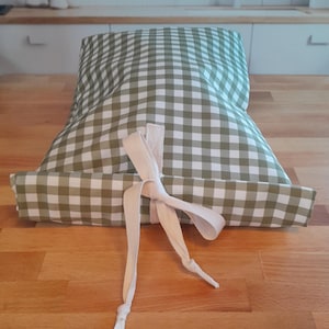 May include: A green and white gingham patterned pillow with a cream-colored ribbon tied in a bow. The pillow is rectangular and appears to be made of fabric. The background is a wooden surface.