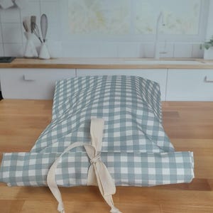 May include: A folded, light blue and white checkered fabric item, tied with a cream-colored ribbon. The item is resting on a wooden surface, with a kitchen setting in the background.