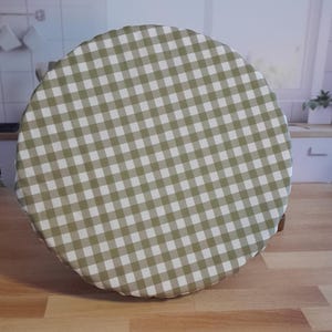 May include: A round, green and white checkered fabric bowl cover. The cover is placed over a wooden bowl, with the checkered pattern visible. The cover is made of a soft, flexible material.
