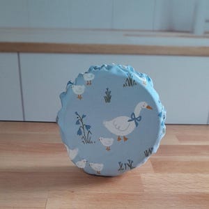 May include: A light blue fabric bowl cover with a charming goose and gosling pattern. The cover has a ruffled edge and is decorated with white geese, small goslings, and floral accents. The cover is designed to fit over a bowl.