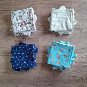 Pode incluir: Quatro pilhas de panos de tecido quadrados com diferentes padrões. Uma pilha tem um fundo branco com um desenho de rato e bagas. Outra tem um padrão de folhas azuis. A terceira tem um fundo azul claro com animais da floresta. A última tem um padrão de bolinhas verde claro.