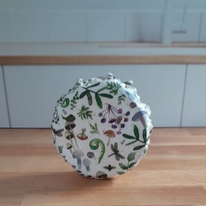 May include: A round, white ceramic jar with a lid, decorated with a botanical pattern of green leaves, mushrooms, and insects. The jar sits on a wooden surface, suggesting a kitchen or dining setting.