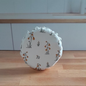 May include: A round, white fabric object with a floral pattern featuring orange and brown flowers and green grass. The object is sitting on a wooden surface. The floral design is repeated across the surface.
