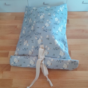 Könnte beinhalten: Hellblaues, rechteckiges Kissen mit einem sich wiederholenden Muster aus weißen Gänsen und grünen Zweigen. Das Kissen ist mit einem cremefarbenen Band gebunden. Das Kissen liegt auf einer Holzoberfläche. Ca. 40 cm lang.