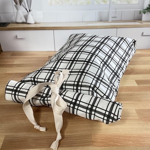 Puede incluir: Un artículo de cocina rectangular enrollado, con un patrón de cuadros blanco y negro, atado con una cinta color crema. El artículo está sobre una superficie de madera, sugiriendo un entorno de cocina. El diseño es simple y moderno.