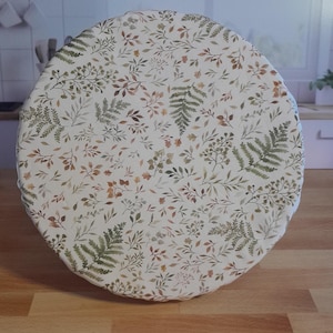 以下が含まれることがあります： オフホワイトの生地で作られた丸い食品カバー。植物柄のデザインで、緑、茶色、ベージュの葉や小枝が散りばめられています。カバーは木製の表面に置かれています。