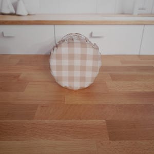 May include: A round jar cover with a beige and white gingham pattern and a frilled edge. It rests on a wooden surface. The background shows a white kitchen with a wooden worktop and a window.