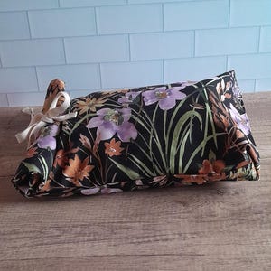 Puede incluir: Una bolsa con estampado floral negro con flores moradas, naranjas y amarillas y hojas verdes. La bolsa está atada con una cinta color crema. La bolsa está sobre una superficie de madera.