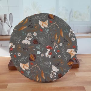 May include: A round, gray fabric bowl cover with a whimsical botanical pattern. The design includes mushrooms, flowers, and leaves in shades of white, orange, and brown. The cover is placed on a wooden tray.
