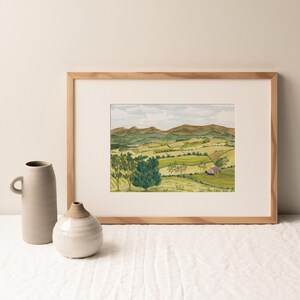 May include: A watercolour painting depicting a rural landscape with green hills, trees, and a red building. The artwork is framed in a light wood frame. Two ceramic vases are in the foreground, adding to the scene.