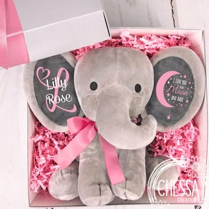 May include: A gray plush elephant stuffed animal with pink ribbon and ears. The ears have black felt circles with pink text. One ear says 'Lilly Rose' and the other ear says 'I love you to the Moon and Back'. The elephant is in a white gift box with pink tissue paper.