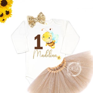 Puede incluir: Un body blanco de manga larga con un número uno marrón y una ilustración de abeja. El body tiene el nombre "Madeline" escrito en purpurina dorada. Un lazo de purpurina dorada está en el body. Una falda de tul beige con purpurina dorada está incluida.